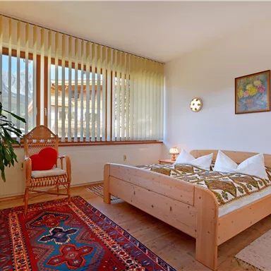 Ein gemütliches Schlafzimmer mit einem großen Bett, bunten Kissen und einem schönen Teppich. Ein Pflanzenständer und große Fenster sorgen für viel Licht.