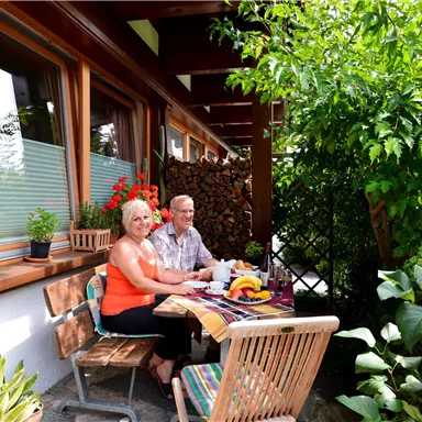 Ein gemütlicher Außenbereich mit zwei Personen, die an einem Tisch mit Früchten sitzen. Umgeben von grünen Pflanzen und Blumen.