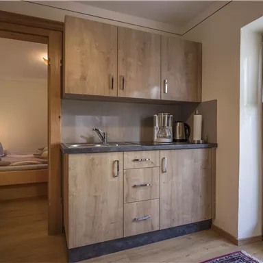 A cozy kitchenette with wooden cabinets and a small sink. In the background, a bedroom with beds and a window can be seen.