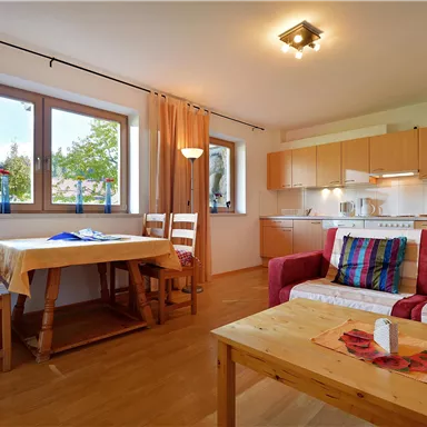 A bright living room with an open kitchen. There is a table with chairs and a cozy seating area with colorful cushions.