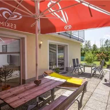 A cozy terrace with a large table and chairs under a red parasol. In the background, green plants and a sunny sky can be seen.