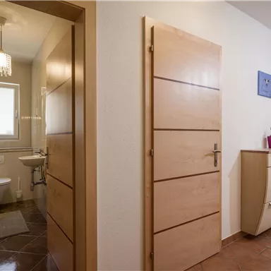 A bright hallway with two closed doors and a modern wardrobe. On the left side, there is a bathroom with a toilet and a window.
