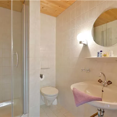 Ein modernes Badezimmer mit einer Dusche, einem Waschbecken und einem großen Spiegel. Die Wände sind hell gefliest und der Boden ist schlicht.