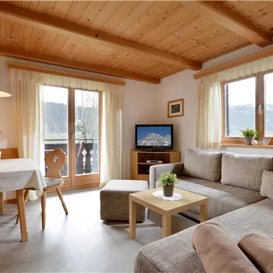 A cozy living room with a wooden ceiling and large windows. There is a sofa, a table, and a television, as well as a view of the mountains.