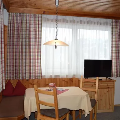 Ein gemütlicher Raum mit einem Esstisch und Stühlen. Große Fenster lassen viel Licht herein und ein Fernseher ist in der Ecke platziert.