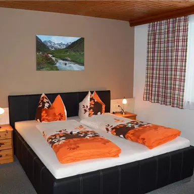 Ein gemütliches Schlafzimmer mit einem Doppelbett, dekoriert in orange und weiß. An der Wand hängt ein Gemälde von einer Berglandschaft und ein Fenster mit Vorhängen lässt natürliches Licht herein.