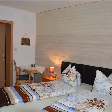 Ein helles Schlafzimmer mit zwei Einzelbetten und bunten Bettbezügen. Es gibt einen kleinen Tisch und einen Stuhl neben der Tür.