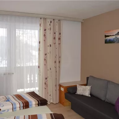 Ein gemütliches Zimmer mit zwei Betten und einem grauen Sofa. Große Fenster mit Vorhängen sorgen für viel Tageslicht.