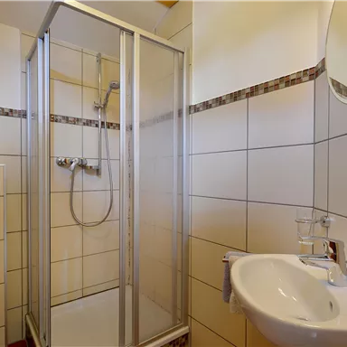 Ein modernes Badezimmer mit einer Duschkabine, einem Waschbecken und einem Spiegel. Die Wände sind hell gefliest und die Ausstattung ist sauber und funktional.