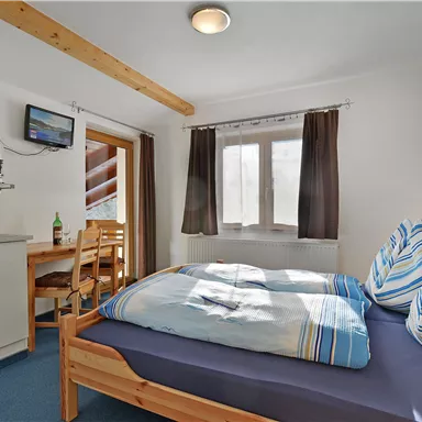 Ein gemütliches Zimmer mit einem Doppelbett und bunten Kissen. Es gibt einen kleinen Esstisch, eine Küchenzeile und ein Fenster mit Blick nach draußen.