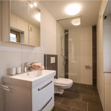 Ein modernes Badezimmer mit hellen Fliesen und einem großen Spiegel. Die Ausstattung umfasst ein Waschbecken, eine Dusche und ein Fenster für Tageslicht.