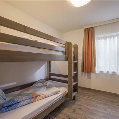 Ein einfaches Zimmer mit einem Hochbett und zwei bequemen Kissen. Das Fenster lässt viel Tageslicht herein, und der Boden ist aus Holz.