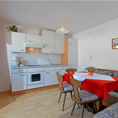 Eine moderne Küche mit einem Essbereich und einem roten Tischdecken. Helle Wände und Fenster sorgen für ein freundliches Ambiente.