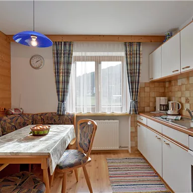 Eine gemütliche Küche mit einem Essbereich und einem Fenster, durch das Tageslicht strömt. Die Holzwände und die farbenfrohen Vorhänge schaffen eine einladende Atmosphäre.