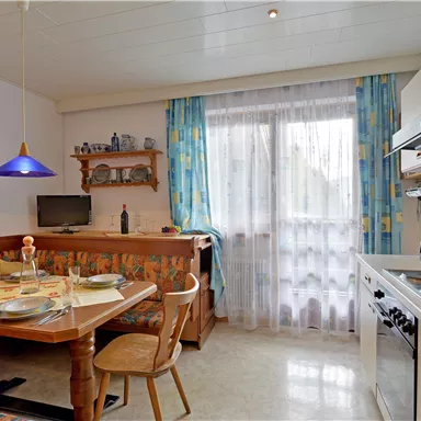 Eine helle Küche mit einem Esstisch und Stühlen. Es gibt Fenster mit hellen Vorhängen und moderne Küchengeräte.