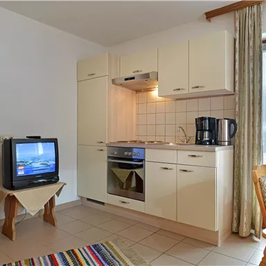 Eine kleine Küche mit einem Herd, Kühlschrank und Kaffeemaschine. Im Hintergrund steht ein Fernseher und ein Tisch mit Stühlen.