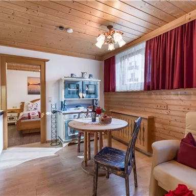 A cozy living kitchen with wooden walls and a round table. In the background, a bedroom and decorative furniture can be seen.