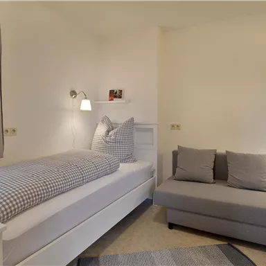 Ein einfaches Schlafzimmer mit einem Einzelbett und einem grauen Sofa. Die Wände sind hell, und es gibt Vorhänge an einem Fenster.