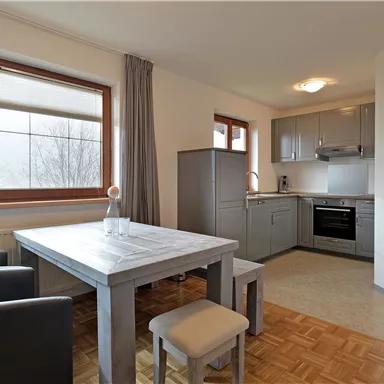 Eine moderne Küchenecke mit einem Tisch und Stühlen. Die Wände sind hell und die Fenster bieten viel Tageslicht.