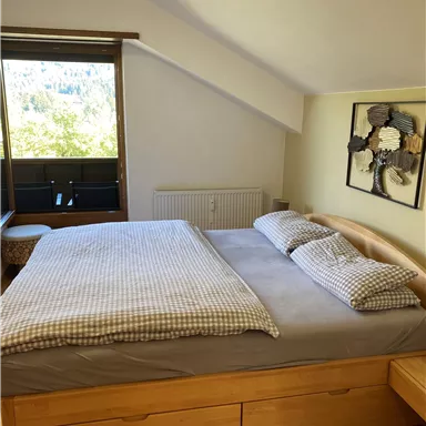 A cozy bedroom with a large bed and pillows. The room has a window overlooking the outdoors and hardwood flooring.