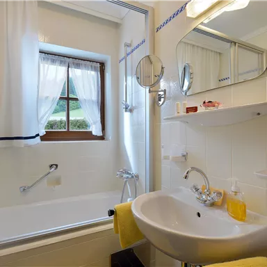 Ein helles Badezimmer mit einer Badewanne und einem Waschbecken. Es gibt Fenster mit Vorhängen und eine große Wandspiegel.