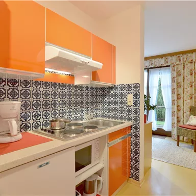 Eine moderne Küche mit orangen Schränken und einer blauen Fliesenwand. Im Hintergrund ist ein gemütlicher Wohnbereich mit einem Sessel und Vorhängen zu sehen.