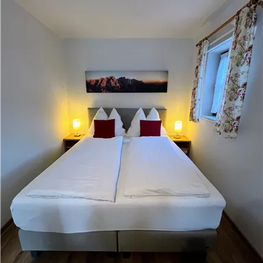 Ein gemütliches Zimmer mit Doppelbett und Nachttischlampen. Das Fenster hat eine florale Gardine und an der Wand hängt ein Bild.
