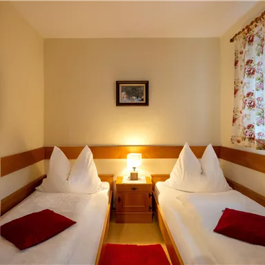 Ein gemütliches Zimmer mit zwei Einzelbetten und einer Nachttischlampe. Das Fenster hat eine florale Gardine und an der Wand hängt ein Bild.