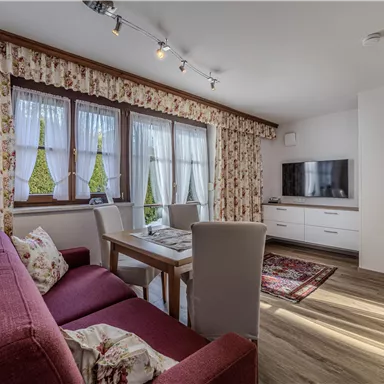 Ein gemütlicher Wohnbereich mit einem roten Sofa und einem Holztisch. Große Fenster lassen viel Licht herein und sorgen für eine einladende Atmosphäre.