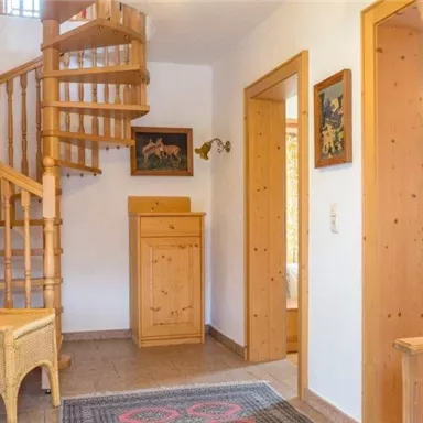 A cozy hallway with a spiral wooden staircase. Pictures hang on the walls, and there is a wicker chair and a dresser.