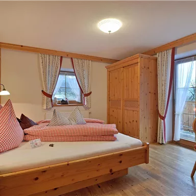 A cozy bedroom with wooden details, two beds, and traditional red pillows. Large windows let in plenty of light and offer a view outside.
