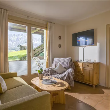 Ein gemütliches Wohnzimmer mit einem grünen Sofa und einem Holztisch. Große Fenster bieten einen Ausblick auf die Natur und die Berge.