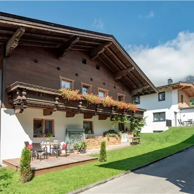 Ein charmantes Holzhaus in den Bergen mit blühenden Balkonpflanzen. Im Hintergrund sind schneebedeckte Gipfel und ein klarer Himmel sichtbar.