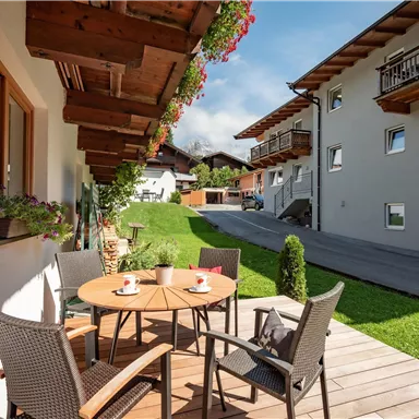 Eine einladende Terrasse mit einem Tisch und Stühlen, umgeben von grünem Rasen. Im Hintergrund sind moderne Gebäude und eine sonnige Landschaft zu sehen.