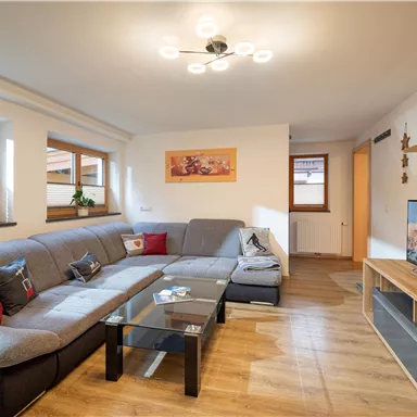 Ein gemütlicher Wohnraum mit einer großen grauen Couch und einem Fernseher. Große Fenster lassen viel Licht herein und schaffen eine einladende Atmosphäre.