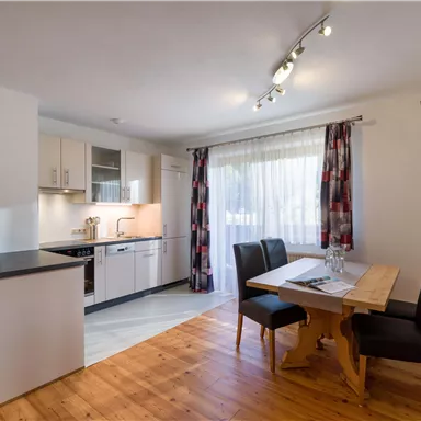 Eine moderne Küche mit hellen Schränken und einem Esstisch. Die großen Fenster lassen viel Tageslicht herein.