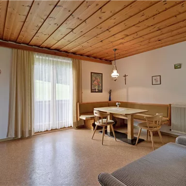 Ein gemütlicher Wohnraum mit Holzdecke und einem Esstisch. Die Fenster lassen viel Tageslicht herein und die Einrichtung ist schlicht und einladend.