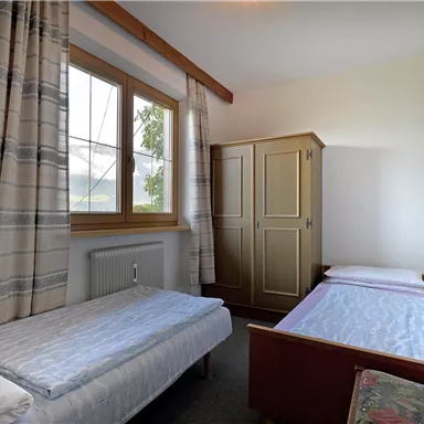 Ein einfaches Schlafzimmer mit zwei Einzelbetten und einem Kleiderschrank. Große Fenster lassen viel natürliches Licht herein.