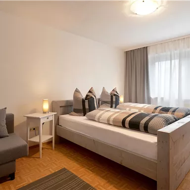 Ein modernes Schlafzimmer mit einem großen Bett und gemütlichen Kissen. Es gibt einen Sitzbereich und viel Tageslicht durch die Fenster.