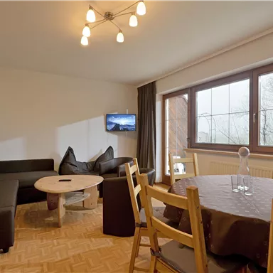 Ein modernes Wohnzimmer mit einem gemütlichen Sofa und einem runden Tisch. Große Fenster bieten einen Blick auf die Außenansicht und lassen viel Tageslicht herein.