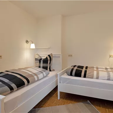 Ein gemütliches Schlafzimmer mit zwei Einzelbetten und modernen Leseleuchten. Die Wände sind neutral, und es gibt dekorative Kissen mit karomuster.