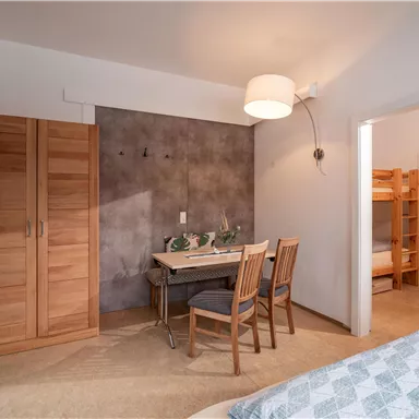 Ein gemütliches Zimmer mit einem Tisch, Stühlen und einem Kleiderschrank aus Holz. Auf der linken Seite ist ein offenes Zimmer mit Etagenbetten sichtbar.