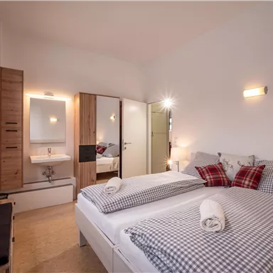 Ein gemütliches Schlafzimmer mit zwei Betten und karierten Bettdecken. Es gibt einen Spiegel, einen kleinen Waschbereich und eine warme Beleuchtung.