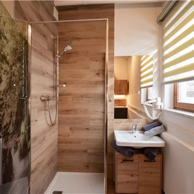 Ein modernes Badezimmer mit einer Dusche und einer Holzverkleidung. Es gibt ein Waschbecken und große Fenster mit Jalousien, die für viel Licht sorgen.