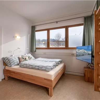 Ein modernes Schlafzimmer mit einem Doppelbett aus Holz und einem Fernseher. Große Fenster bieten Blick auf die Landschaft und lassen viel Licht herein.