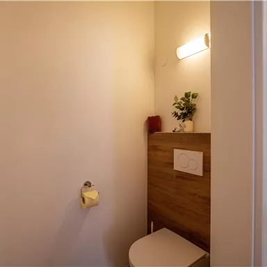 Ein kleines, modernes Badezimmer mit einer weißen Toilettenanlage und einer Holzablage. Die Wände sind hell und eine Pflanze sowie ein Handtuch verleihen dem Raum eine persönliche Note.