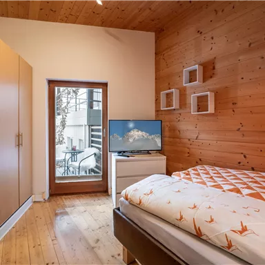 Ein gemütliches Schlafzimmer mit Holzvertäfelung, einem Fernseher und einem Fenster mit Blick auf einen Balkon. Die Einrichtung ist modern und einladend.