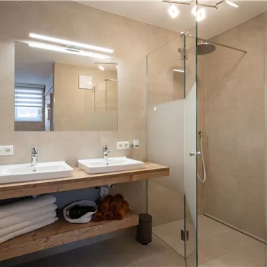 Ein modernes Badezimmer mit Doppelwaschbecken und einer Glasdusche. Die Wände sind in neutralen Tönen gehalten, und im Hintergrund sind gefaltete Handtücher sichtbar.