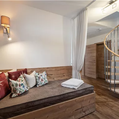 Ein gemütlicher Raum mit einer Holzbank und bunten Kissen. Eine geschwungene Treppe führt in die nächste Etage.