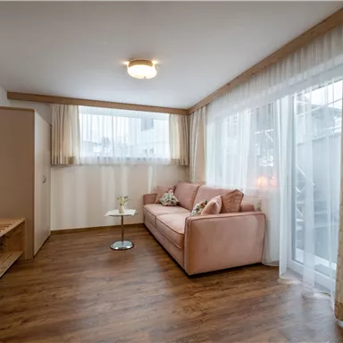 Ein gemütliches Wohnzimmer mit einem rosa Sofa und einem kleinen Tisch. Große Fenster sorgen für viel natürliches Licht und eine freundliche Atmosphäre.